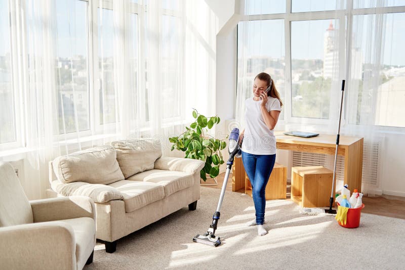 Young woman in white shirt holding vacuum cleaner and talking on cellphone, copy space. Housework, spring-cleaning