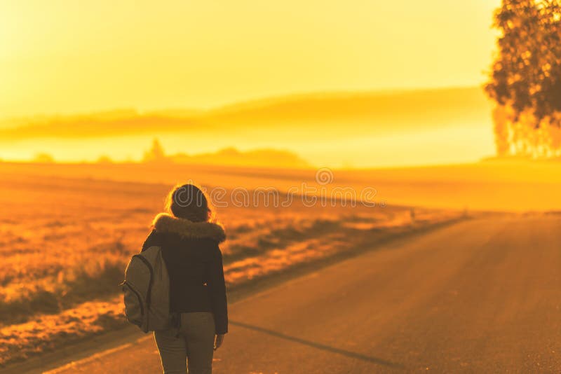 Young woman in the winter jacket with a backpack on the road against the background of the sunrise autumn field.