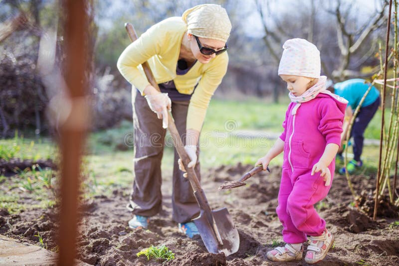 Young woman working in garden while her little daughter playing with spade beside her