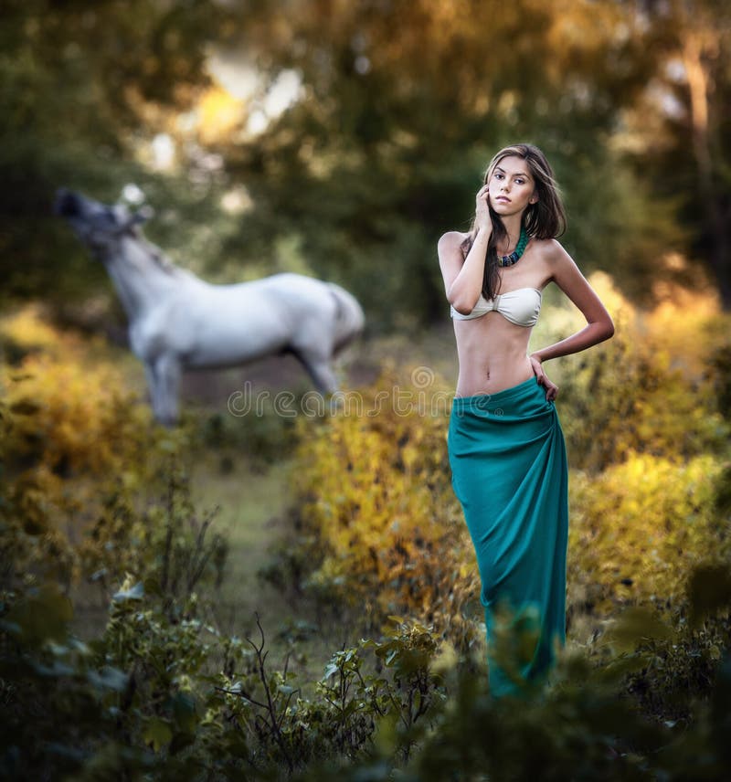 Young woman in a blue long skirt and white bra at sunset in forest with a white horse in background .Beautiful young woman with long hair in garden with wild horse. Girl and horse in the field. Young woman in a blue long skirt and white bra at sunset in forest with a white horse in background .Beautiful young woman with long hair in garden with wild horse. Girl and horse in the field