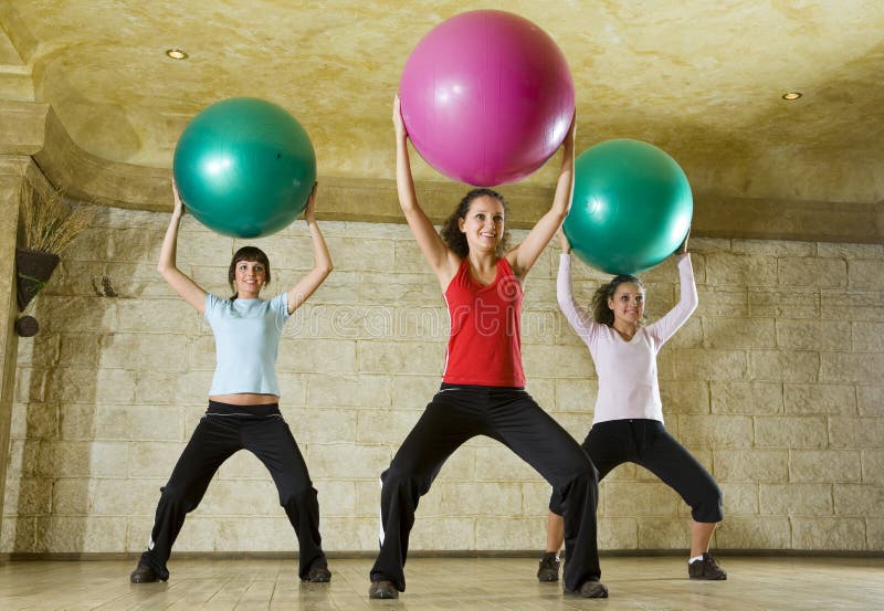 A group of women making exercise holding big balls over heads and standing astride. They're smiling. Low angle view. A group of women making exercise holding big balls over heads and standing astride. They're smiling. Low angle view.