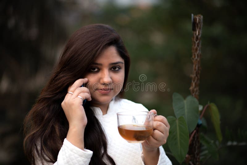 Young woman holding a transparent tea cup with black tea in a garden wearing white woollen jacket. Young woman holding a transparent tea cup with black tea in a garden wearing white woollen jacket