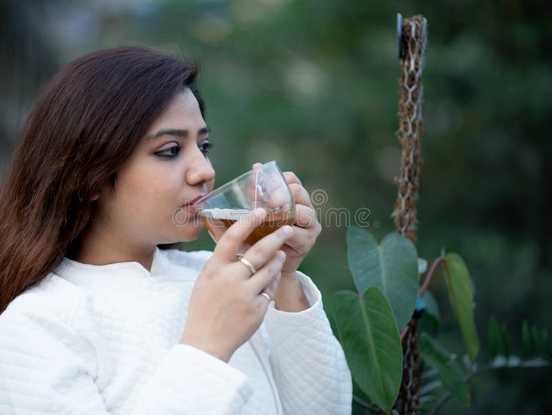 Young woman holding a transparent tea cup with both hands sipping black tea in a garden wearing white woollen jacket. Young woman holding a transparent tea cup with both hands sipping black tea in a garden wearing white woollen jacket