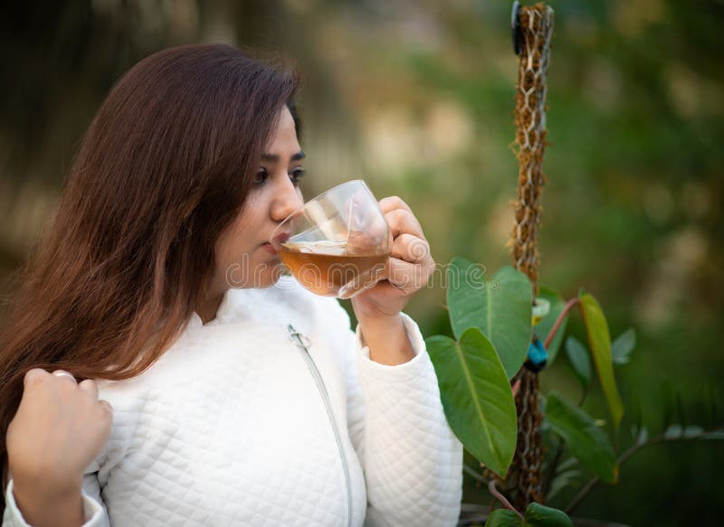 Young woman holding a transparent tea cup sipping black tea in a garden wearing white woollen jacket. Young woman holding a transparent tea cup sipping black tea in a garden wearing white woollen jacket