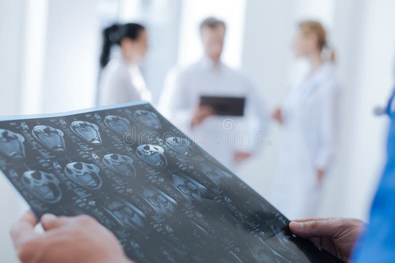 Diagnosing urgent health condition. Attentive experienced concentrated radiologic technologist working at the medical lab and examining the ct scan while his colleagues using tablet in the background. Diagnosing urgent health condition. Attentive experienced concentrated radiologic technologist working at the medical lab and examining the ct scan while his colleagues using tablet in the background