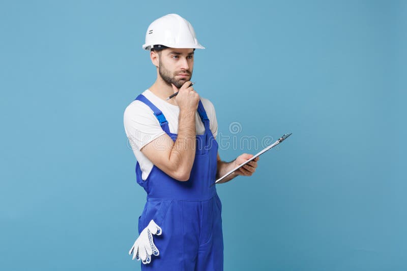 Pensive young man in coveralls protective helmet hardhat hold clipboard with papers document isolated on blue background. Instruments accessories for renovation apartment room. Repair home concept. Pensive young man in coveralls protective helmet hardhat hold clipboard with papers document isolated on blue background. Instruments accessories for renovation apartment room. Repair home concept