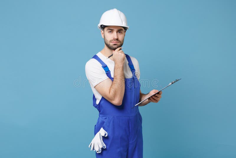 Pensive young man in coveralls protective helmet hardhat hold clipboard with papers document isolated on blue background. Instruments accessories for renovation apartment room. Repair home concept. Pensive young man in coveralls protective helmet hardhat hold clipboard with papers document isolated on blue background. Instruments accessories for renovation apartment room. Repair home concept