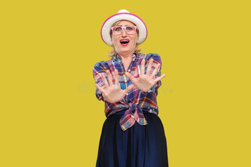 Please stop, i don`t need this. Portrait of scared modern stylish mature woman in casual style with hat and eyeglasses standing with blocking hands and panic. studio shot isolated on yellow background. Please stop, i don`t need this. Portrait of scared modern stylish mature woman in casual style with hat and eyeglasses standing with blocking hands and panic. studio shot isolated on yellow background