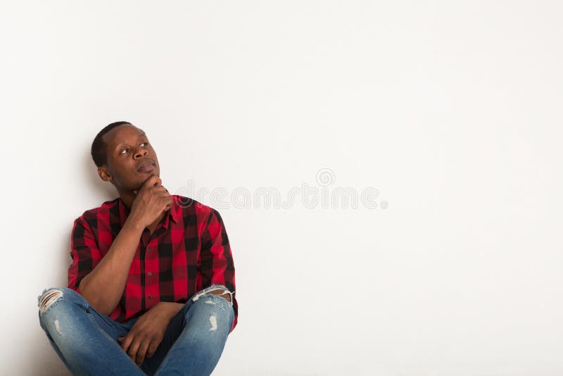 Thinking, pensive young black man sits leaning on the wall in casual looking aside at white studio background. Boy in checkered shirt sitting with crossed legs. Crop, close up, copy space. Thinking, pensive young black man sits leaning on the wall in casual looking aside at white studio background. Boy in checkered shirt sitting with crossed legs. Crop, close up, copy space
