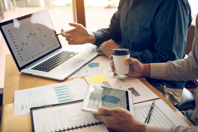 Business partnership pointing to the graph of the company financial statements report and profits earned during in the computer screen with giving a presentation to colleagues holding tablet and coffee in office room. Business partnership pointing to the graph of the company financial statements report and profits earned during in the computer screen with giving a presentation to colleagues holding tablet and coffee in office room.