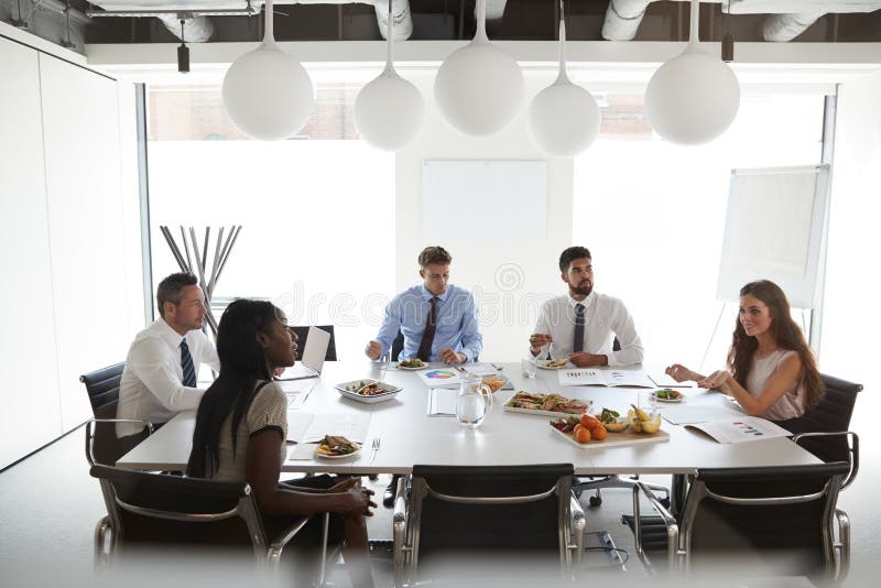 Zakenlieden en Onderneemsters die in Moderne Bestuurskamer bij Werkende Lunch samenkomen stock afbeelding Zakenlieden en Onderneemsters die in Moderne Bestuurskamer bij Werkende Lunch samenkomen stock afbeelding
