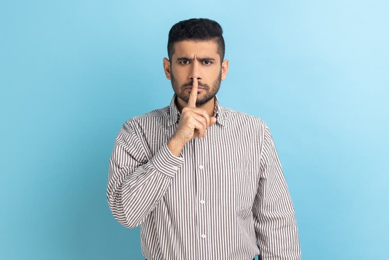 Please, be quiet. Businessman holding finger on lips making hush silence gesture, asking to keep secret, don& x27;t speak, wearing striped shirt. Indoor studio shot isolated on blue background. Please, be quiet. Businessman holding finger on lips making hush silence gesture, asking to keep secret, don& x27;t speak, wearing striped shirt. Indoor studio shot isolated on blue background.