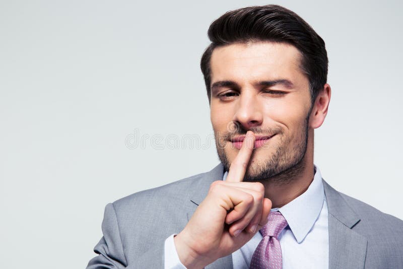 Businessman pointing finger over lips, asking for silence over gray background. Looking at camera. Businessman pointing finger over lips, asking for silence over gray background. Looking at camera