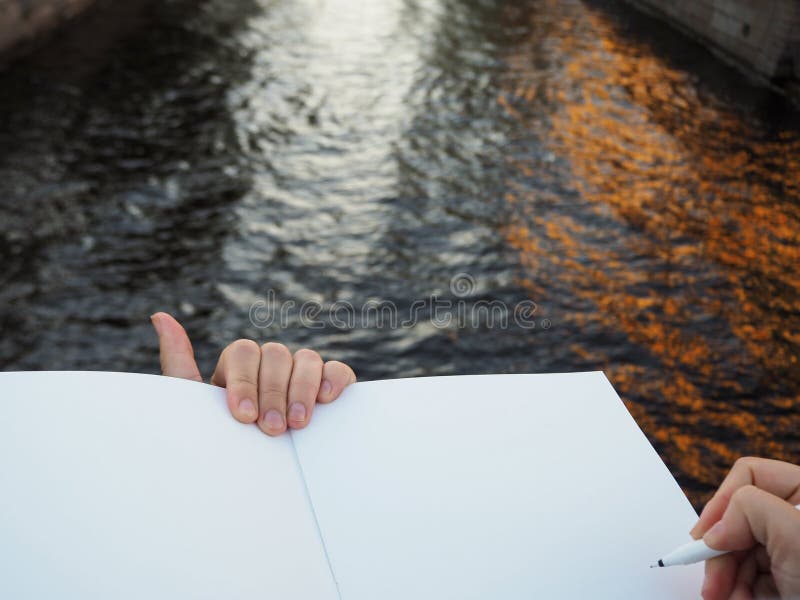Zakończenie w górę żeńskiej ręki trzyma rozpieczętowane sketchbook strony na miasto kanału tle i pióro zdjęcia stock Zakończenie w górę żeńskiej ręki trzyma rozpieczętowane sketchbook strony na miasto kanału tle i pióro zdjęcia stock
