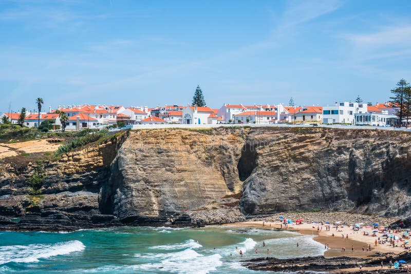 Houses on a cliff by the sea in the fishing village of Zambujeira do Mar in the municipality of Odemira. Several people enjoying the ocean waves on the beach in summer. Beach known for water sports like surfing and paddle. Portugal. Houses on a cliff by the sea in the fishing village of Zambujeira do Mar in the municipality of Odemira. Several people enjoying the ocean waves on the beach in summer. Beach known for water sports like surfing and paddle. Portugal