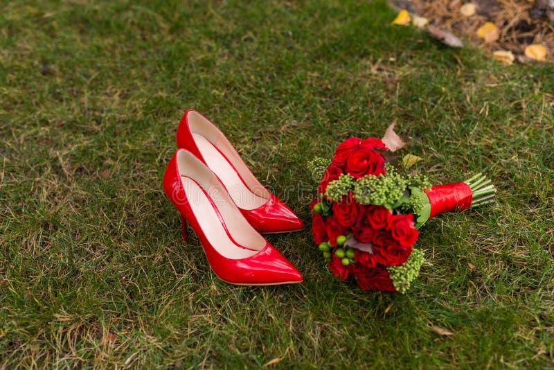 Zapatos rojos de la boda de la moda femenina con el ramo del ` s de la novia de rosas rojas y de bayas verdes en fondo de la hier imagenes de archivo Zapatos rojos de la boda de la moda femenina con el ramo del ` s de la novia de rosas rojas y de bayas verdes en fondo de la hier imagenes de archivo