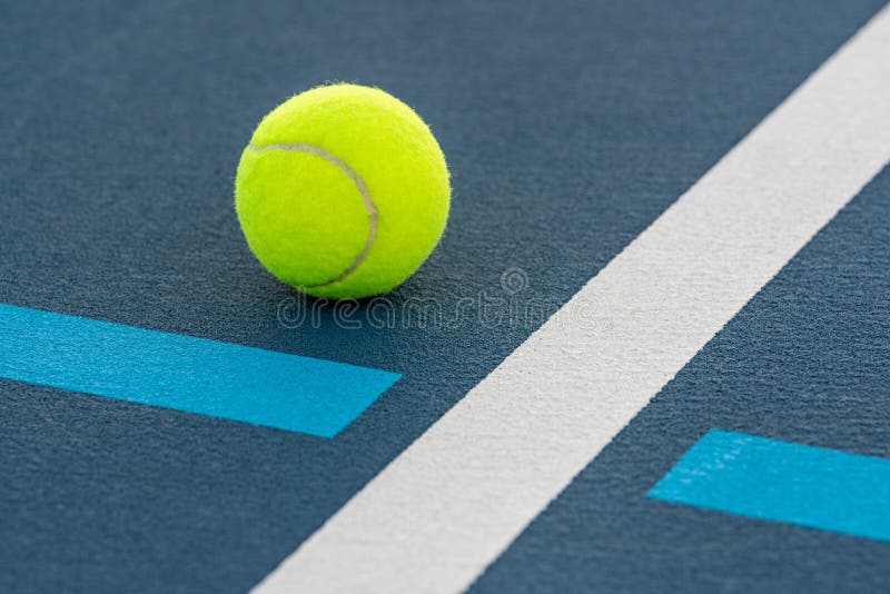 A closeup of a tennis ball at the intersection of blue tennis courts with white and light blue lines. A closeup of a tennis ball at the intersection of blue tennis courts with white and light blue lines