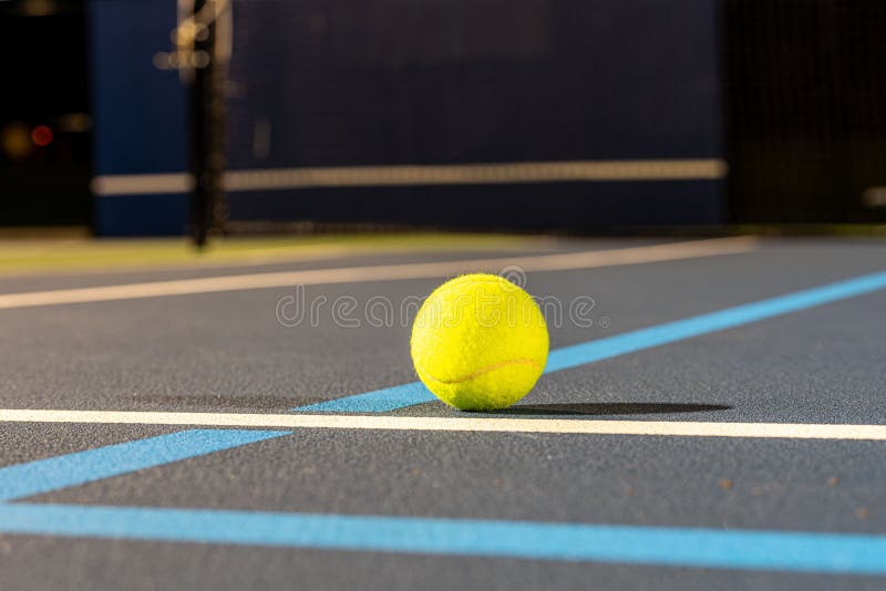 A closeup of a tennis ball at the intersection of blue tennis courts with white and light blue lines. A closeup of a tennis ball at the intersection of blue tennis courts with white and light blue lines