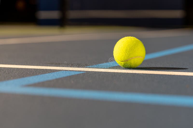 A closeup of a tennis ball at the intersection of blue tennis courts with white and light blue lines. A closeup of a tennis ball at the intersection of blue tennis courts with white and light blue lines