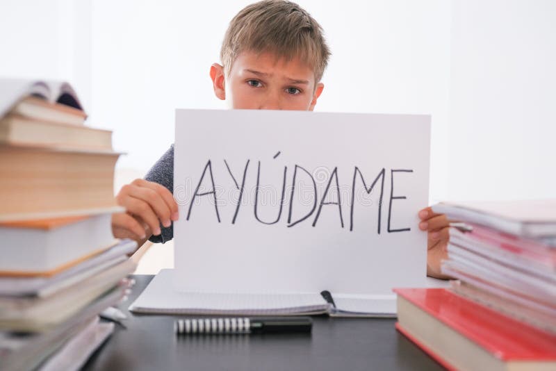 Tired boy sitting at the table with many books, exercises books. Spanish word Auydame - Help me- is written on sheet of paper. Learning difficulties, school, quarantine education concept. Tired boy sitting at the table with many books, exercises books. Spanish word Auydame - Help me- is written on sheet of paper. Learning difficulties, school, quarantine education concept.