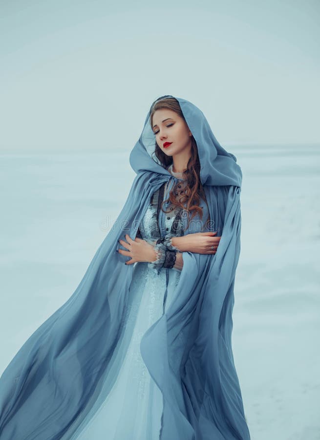 Art photo. Fantasy young woman fairy elf in blue cape with hood stands in cold wind. Winter nature background, white snow. Girl Queen walks in medieval dress, silk cloak, fabric is waving, fluttering. Art photo. Fantasy young woman fairy elf in blue cape with hood stands in cold wind. Winter nature background, white snow. Girl Queen walks in medieval dress, silk cloak, fabric is waving, fluttering.