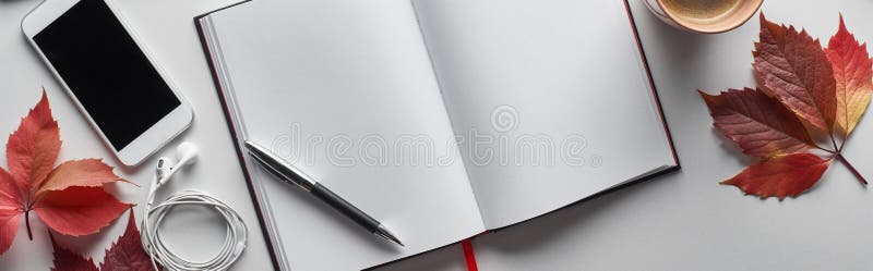 Panoramic shot of blank notebook with pen, smartphone, earphones and red leaves of wild grapes on white table,stock image. Panoramic shot of blank notebook with pen, smartphone, earphones and red leaves of wild grapes on white table,stock image