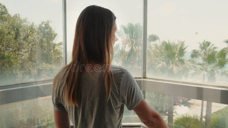 Zdjęcie z tyłu pięknej kobiety, wznoszącej się w przezroczystej windzie, oglądającej zmianę widoku z roślin tropikalnych na morze.