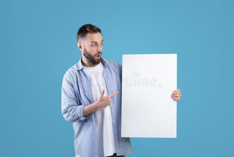 Zdziwiony tysiącletni mężczyzna wskazujący biały pusty plakat na niebieskim tle studio makijaż dla Twojego projektu reklamy obraz stock