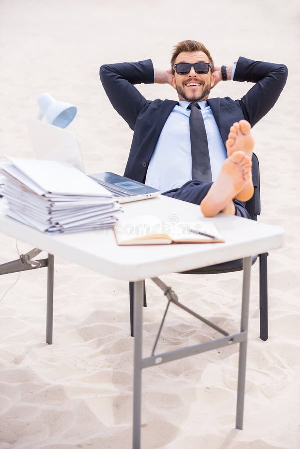 Cheerful young man in formalwear and sunglasses holding hands behind head and holding his feet on the table standing on sand. Cheerful young man in formalwear and sunglasses holding hands behind head and holding his feet on the table standing on sand