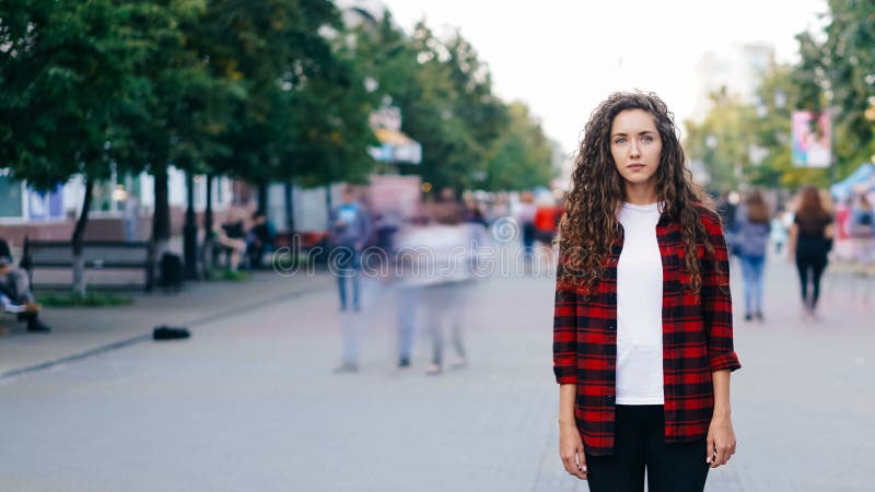 Zeit-Versehenporträt der confedent jungen Frau, die allein in der Straße am Sommertag steht und Kamera während betrachtet