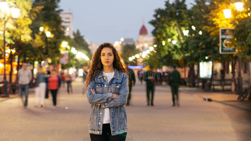 Zeit-Versehenporträt einsamer junger Dame, die in der Straße im Stadtzentrum gelegen mit den Armen gekreuzt steht, während Mengen