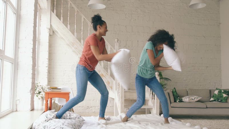 Zeitlupe von zwei jungen hübschen Mädchen der Mischrasse, die zu Hause auf die Bett- und Kampfkissen haben Spaß springen