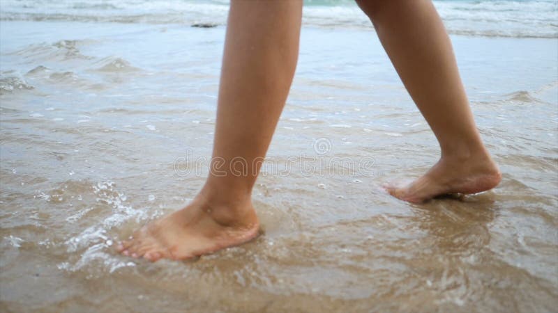 Zeitlupefrauenfüße, die auf den Sandstrand mit Naturseewellenküste für Sommerferienkonzept gehen