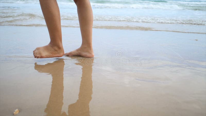 Zeitlupefrauenfüße, die auf den Sandstrand mit Naturseewellenküste für Sommerferienkonzept gehen