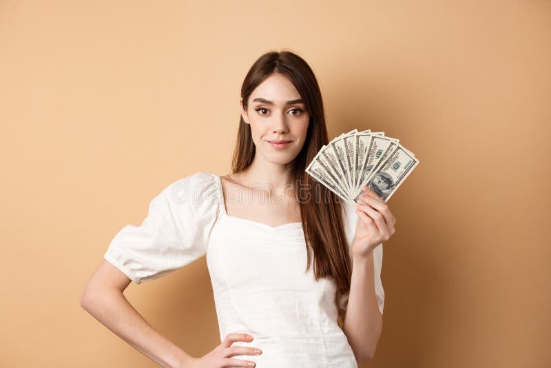 Zelfverzekerde glimlachende vrouw die laat zien dat dollarbiljetten geld verdienen en er tevreden uitzien als ze op beige achtergr