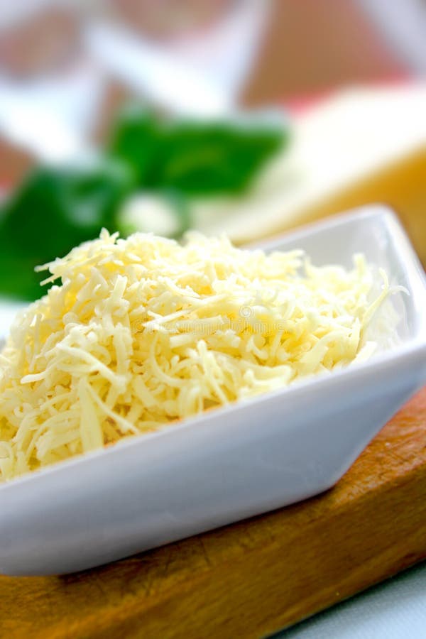 A bowl of grated mature cheddar cheese on wooden chopping board with block of cheese in background and spring of fresh green basil. A bowl of grated mature cheddar cheese on wooden chopping board with block of cheese in background and spring of fresh green basil.