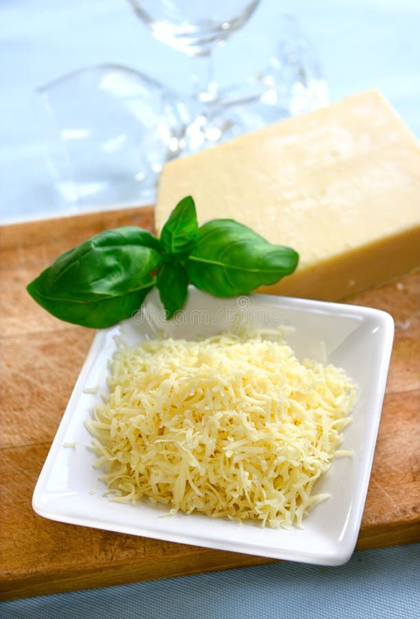 A bowl of grated matture cheddar cheese on wooden chopping board with block of cheese in background and spring of fresh green basil. A bowl of grated matture cheddar cheese on wooden chopping board with block of cheese in background and spring of fresh green basil.