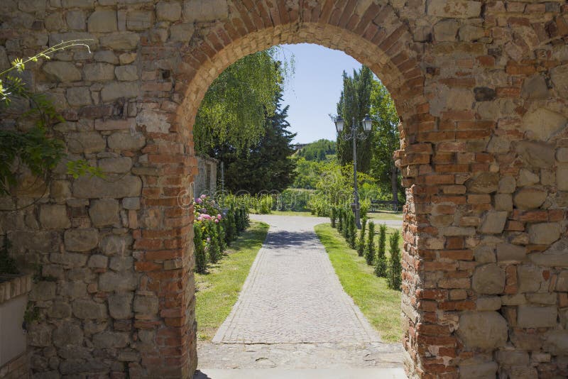 Brickstone arch and passage  in Fortunago, one of the most charming villages of Oltrepò Pavese, Lombardia countryside, Italy. Green park in the background. Brickstone arch and passage  in Fortunago, one of the most charming villages of Oltrepò Pavese, Lombardia countryside, Italy. Green park in the background