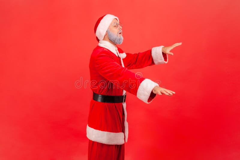 Side view of blind disoriented elderly man with gray beard wearing santa claus costume standing with closed eyes and try to touch something. Indoor studio shot isolated on red background. Side view of blind disoriented elderly man with gray beard wearing santa claus costume standing with closed eyes and try to touch something. Indoor studio shot isolated on red background.