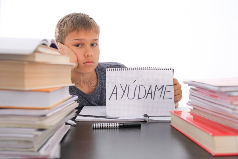 Tired frustrated boy sitting at the table with many books, exercises books. Spanish word Auydame - Help me- is written on open notebook. Learning difficulties, school, quarantine education concept. Tired frustrated boy sitting at the table with many books, exercises books. Spanish word Auydame - Help me- is written on open notebook. Learning difficulties, school, quarantine education concept.