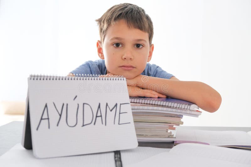 Tired frustrated boy sitting at the table with many books, exercises books. Spanish word Auydame - Help me- is written on open notebook. Learning difficulties, school, quarantine education concept. Tired frustrated boy sitting at the table with many books, exercises books. Spanish word Auydame - Help me- is written on open notebook. Learning difficulties, school, quarantine education concept.