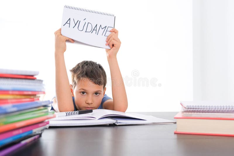 Tired frustrated boy sitting at the table with many books, exercises books. Spanish word Auydame - Help me- is written on open notebook. Learning difficulties, school, education concept. Tired frustrated boy sitting at the table with many books, exercises books. Spanish word Auydame - Help me- is written on open notebook. Learning difficulties, school, education concept.