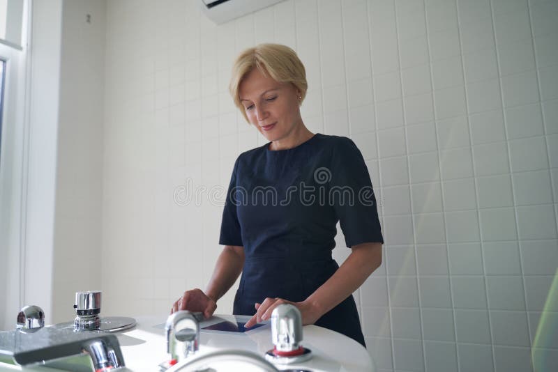 Calm young woman working in a beauty salon and looking at the small monitor while adjusting the settings of a modern bathtub for hydrotherapy. Calm young woman working in a beauty salon and looking at the small monitor while adjusting the settings of a modern bathtub for hydrotherapy