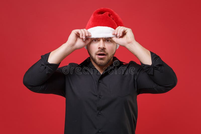 Zszokowany, młody, brodaty mężczyzna z santa w klasycznej, czarnej koszuli świątecznej kapelusz wyizolowany na czerwonym tle w stu