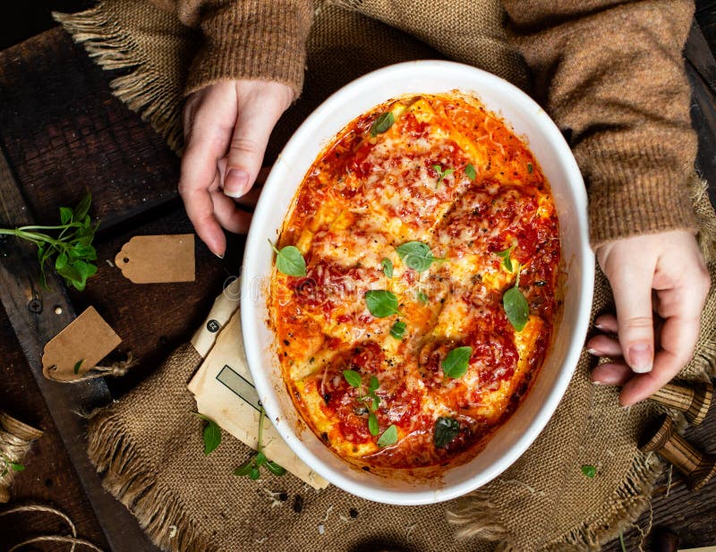 Overhead shot of homemade zucchini ravioli or lasagna in tomato sauce with melted cheese, fresh basil in oval white ceramic baking pan on rustic wooden table. Overhead shot of homemade zucchini ravioli or lasagna in tomato sauce with melted cheese, fresh basil in oval white ceramic baking pan on rustic wooden table