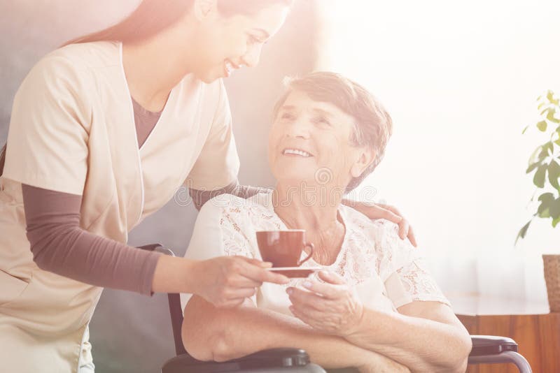 Smiling nurse giving hot tea to happy elderly lady in a wheelchair at nursing home. Smiling nurse giving hot tea to happy elderly lady in a wheelchair at nursing home