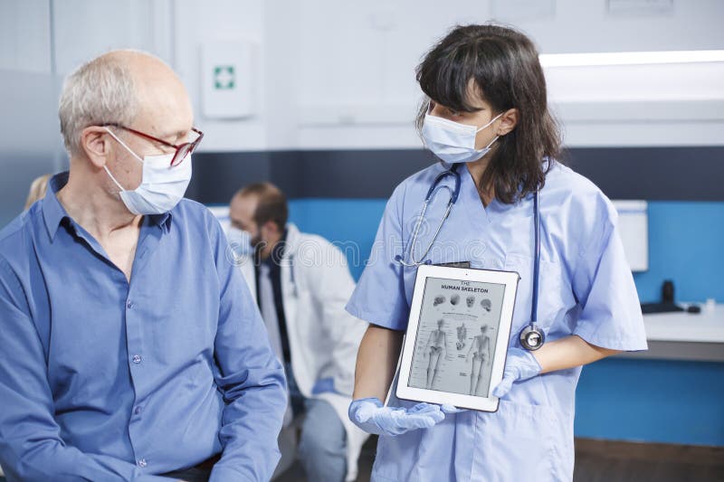 Healthcare specialist describes human skeleton image on tablet to senior patient with face mask at medical exam. Female physician grasps device with osteopathy diagnosis, showing bones examination. Healthcare specialist describes human skeleton image on tablet to senior patient with face mask at medical exam. Female physician grasps device with osteopathy diagnosis, showing bones examination.