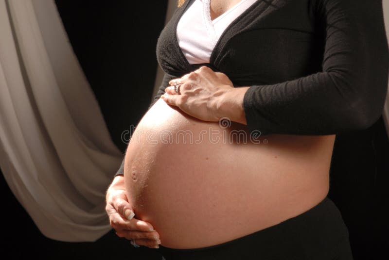 Pregnant woman poses against a black background. Woman is cradling her enlarged stomach with her hands and is dressed in black and white clothing. Woman is nine months pregnant. Pregnant woman poses against a black background. Woman is cradling her enlarged stomach with her hands and is dressed in black and white clothing. Woman is nine months pregnant.