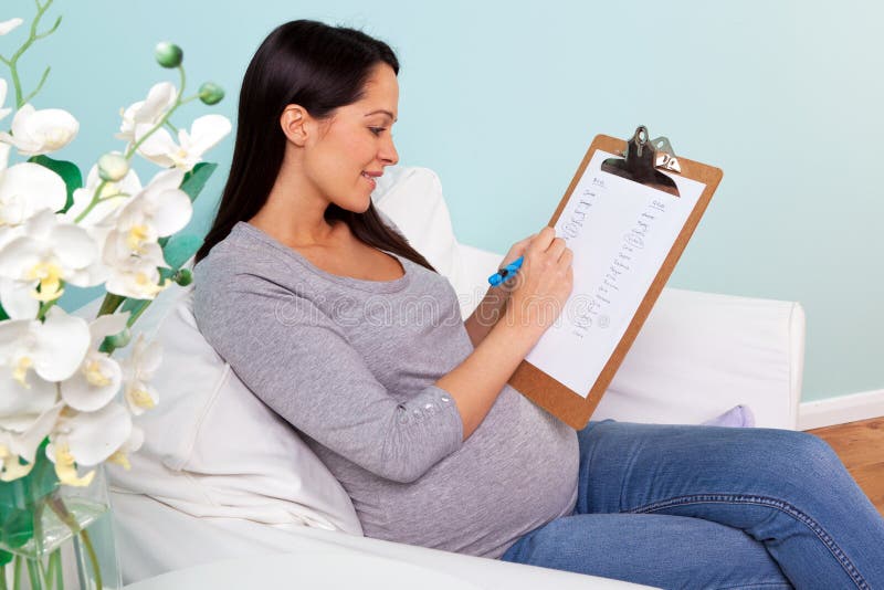 Photo of a pregnant woman at home sitting in an armchair writing a list of possible boy and girl names for her baby. Photo of a pregnant woman at home sitting in an armchair writing a list of possible boy and girl names for her baby.