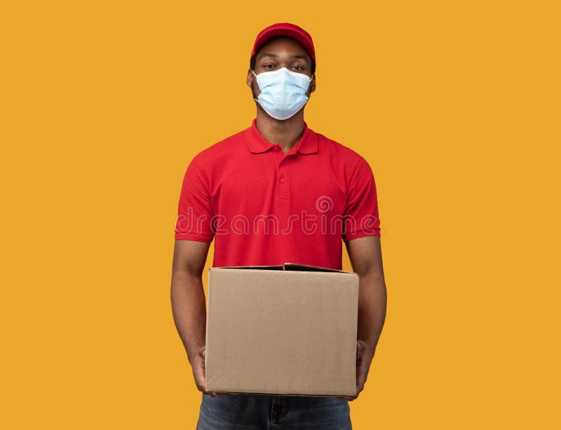 Shopping And Shipping At Quarantine. African american delivery guy in protective face mask and red uniform holding cardboard package isolated over orange studio background. Post during pandemic. Shopping And Shipping At Quarantine. African american delivery guy in protective face mask and red uniform holding cardboard package isolated over orange studio background. Post during pandemic